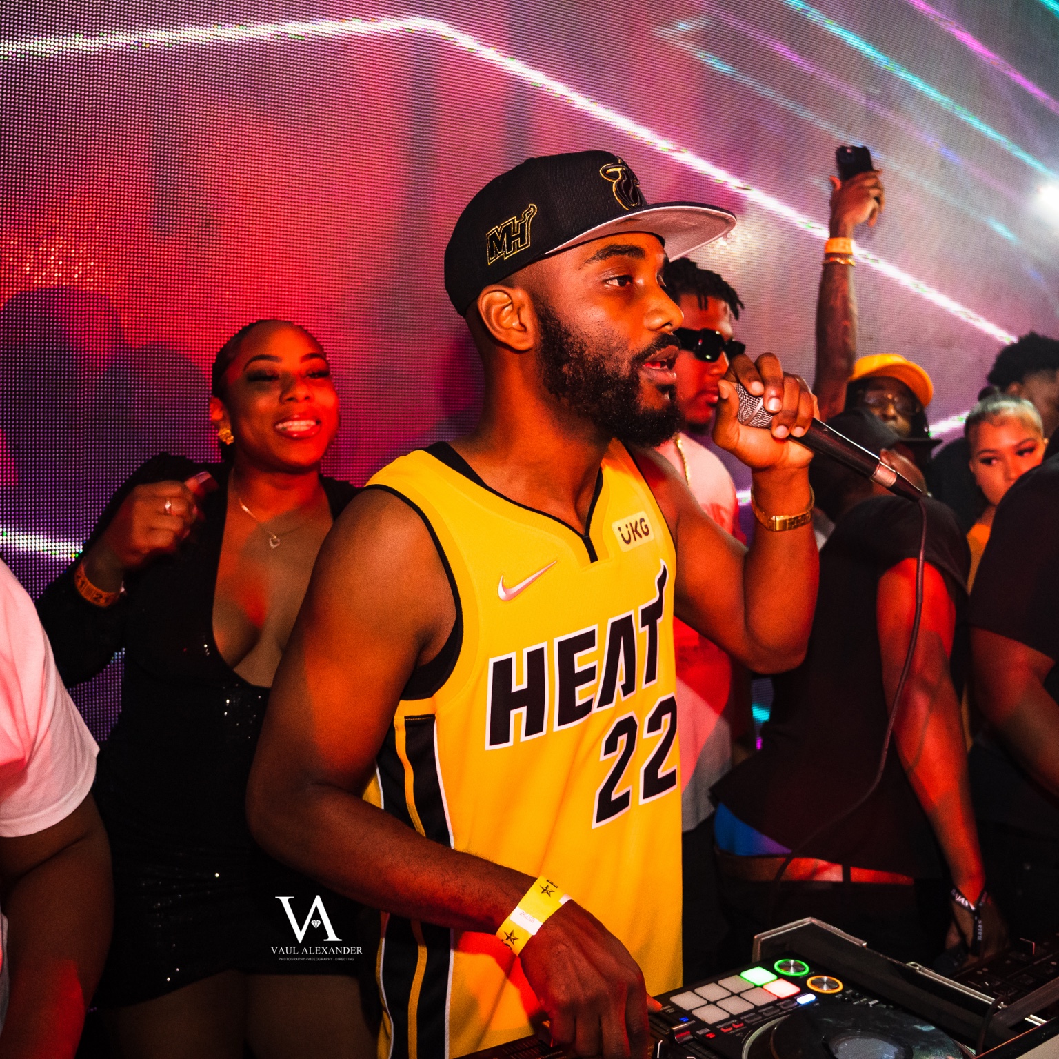 Radio Jamez on the microphone behind the DJ decks in a yellow Miami jersey